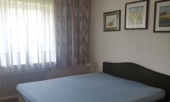 A bedroom with a bed and a nightstand. There is a large window with curtains and some pictures on the wall.