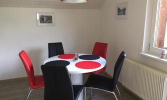 A modern dining area with a round table and colorful chairs. The walls are bright and there is a large window.