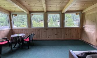 A cozy room with wooden paneling and large windows. There is a table with chairs and a view of the nature outside.