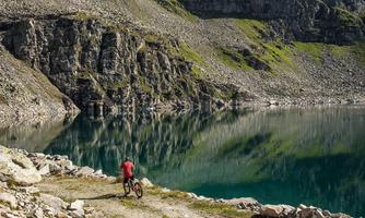 A cyclist stands by the edge of a clear lake, surrounded by steep cliffs and green meadows. The calm water surface reflects the stunning landscape.