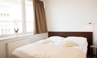 A cozy bedroom with a double bed and white sheets. Next to the window, there is a small table and a plant.
