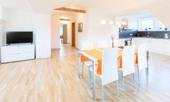 A bright and modern living and dining room with a large dining table and white chairs. The kitchen is open and well-equipped, and the space has a friendly and inviting character.