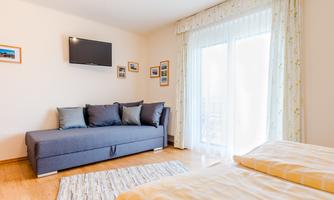 A bright room with a gray sofa and a TV on the wall. Large windows let in a lot of light and create an inviting atmosphere.