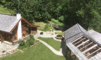 A cozy wooden cabin surrounded by green forest and grass. In the foreground, there is a paved path and a garden area with a fire pit.