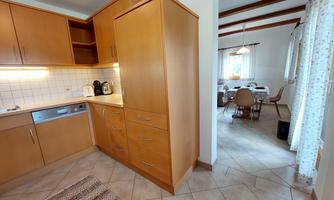 A modern kitchen with wooden cabinets and bright tiles. In the background, there is a dining area with a table and chairs.