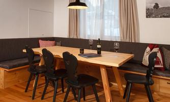 A cozy dining area with a large wooden table and six black chairs. On the table are a bottle of wine and two glasses.