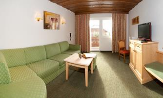 A cozy living room view with a green couch and a table in the middle. A TV and a chair complement the furnishings, with plenty of natural light from the balcony door.