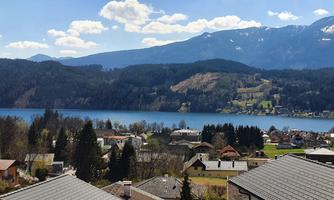 A picturesque view of a lake, surrounded by mountains and idyllic houses. The sky is clear with some clouds.