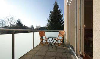 A cozy balcony with two wooden chairs and a small table. In the background, trees and a clear sky can be seen.