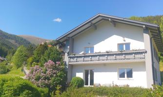 A beautiful house with a balcony, surrounded by green trees and flowers. In the background, mountains and a blue sky can be seen.