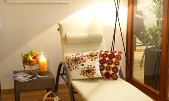 A cozy lounge chair with colorful cushions in a bright room. Next to the chair are a lamp, fruit, and a candle.