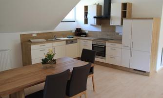 A modern kitchen with wooden and white furniture. At the table are four chairs and a small flower vase.