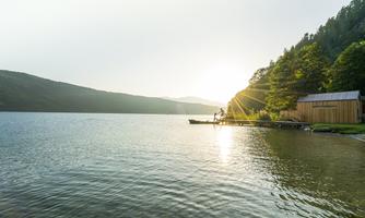 A calm lake with gentle waves and a clearing by the shore. The sun rises behind the mountains, illuminating the scene.