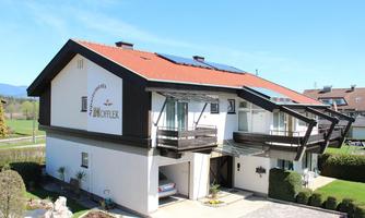 A modern house with a roof made of red tiles and large balconies. In the surrounding area, there are well-maintained green spaces and a clear blue sky.
