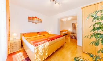 A bright bedroom with a large bed and orange bedding. The room has a warm wooden floor and a friendly atmosphere.