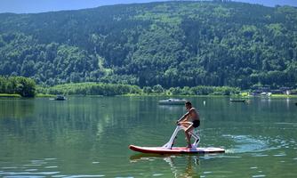 A man is standing on a stand-up paddleboard and paddling across a clear lake. In the background, green hills and trees can be seen.