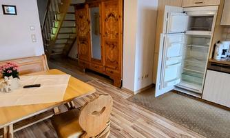 A cozy living space with a dining table and a wooden dresser. In the background, there is an open refrigerator door and a staircase leading up.