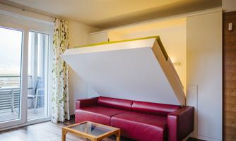 A modern living room with a red sofa and a foldable bed. The room is bright and inviting, with a wooden table.