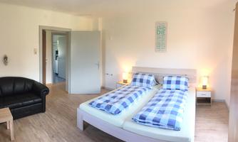 A spacious bedroom with two beds and blue bed covers. There is a cozy sofa and a wooden floor.