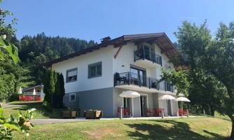 A modern, two-story house surrounded by green landscape. Umbrellas and garden furniture are ready on the terrace.