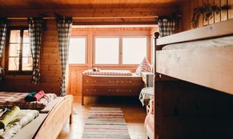 A cozy room with wooden paneling and large windows. There is a bunk bed and a single bed visible, as well as striped curtains.