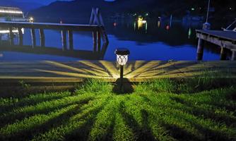 A illuminated garden lamp shines over the grass by the water. In the background, boats and a peaceful nighttime landscape can be seen.