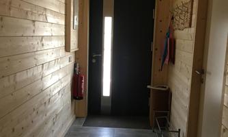 A bright corridor with wooden paneling and a dark door. At the end of the corridor, there is an emergency exit and some pieces of furniture.