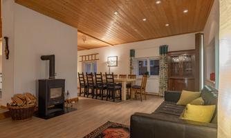 A cozy living room with a black sofa, a table, and chairs. The room features wooden ceilings and a modern fireplace.