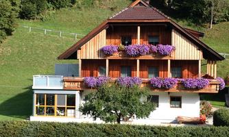 A traditional alpine house with a wooden façade and blooming flowers on the balconies. Surrounded by green meadows and a clear sky.