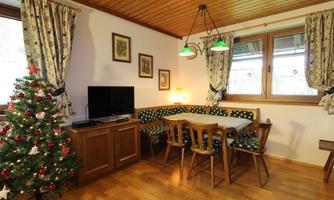 A cozy living room with a Christmas tree and a dining table. The walls are decorated with pictures, and there are large windows with curtains.