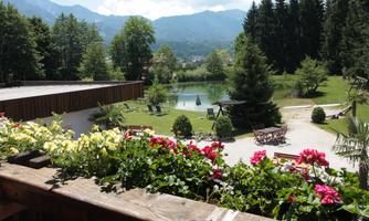 A beautiful garden with a green meadow, a pond, and many trees. Colorful flowers adorn the foreground and the mountains are visible in the background.