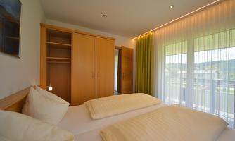 A bright bedroom with two beds and a large window. The room has a modern wardrobe and green curtains.