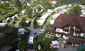A campsite with many motorhomes and green trees. In the background, a lake and a cozy accommodation can be seen.