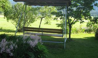 A cozy swing bench is in a green garden. In the background, trees and hills are visible.