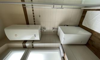 A modern bathroom with a shower, a window, and a heating unit on the wall. Next to the shower is a washing machine.