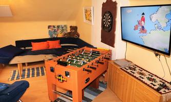 A cozy room with a foosball table, a TV on the wall, and a comfortable couch. The decoration consists of pictures and a large teddy bear.