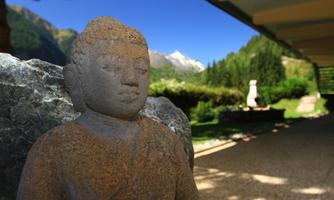 A stone Buddha statue stands in a green environment. In the background, mountains and a clear blue sky are visible.