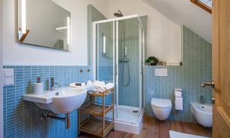 A modern bathroom with blue tiles and a glass shower. There is a sink, a toilet, and a wooden shelf with towels.