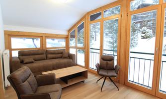 A cozy living room with a brown sofa and two chairs. Large windows offer a view of the snow-covered landscape outside.