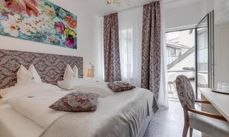 An elegant bedroom with two beds, white bedspreads, and tasteful wall decoration. Large windows provide plenty of daylight and an inviting view outside.