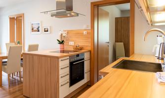 A modern kitchen with wooden surfaces and a cooking island. In the background, a dining area and an entrance door are visible.