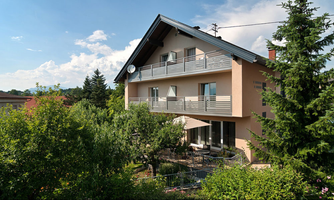 A modern house surrounded by a green garden and trees. The facade is light and features a beautiful balcony.