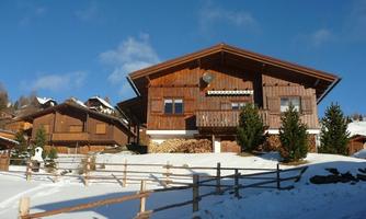 A beautiful wooden house in a snowy landscape. In the background, other chalets and a blue sky can be seen.