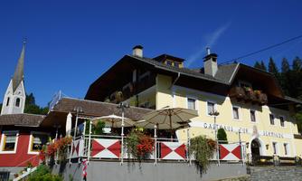 A charming inn with blooming balcony plants and an inviting outdoor area. In the background, a church tower and a beautiful mountain landscape are visible.