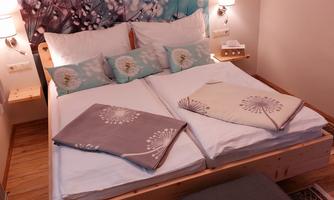 A cozy bedroom with a large bed and colorful wall decor. There are pillows and blankets in soft colors, as well as a small stool.