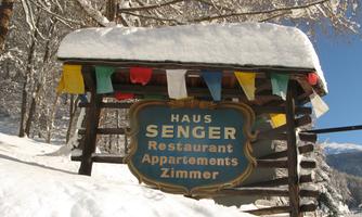 A wooden sign with the inscription "Haus Senger" stands in the snow. It is surrounded by colorful flags and snow-covered trees.