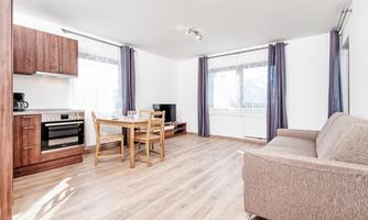 A bright, modern apartment with an open kitchen and a cozy living area. There is a dining table, a television, and large windows that provide plenty of daylight.