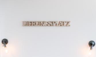 A minimalist room with a white wall background. On the wall, it says "LIEBUNGSPATZ" in wooden letters, flanked by two lamps.