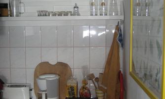 A small kitchen with a shelf holding glasses and a clock. On the countertop, there is a toaster, a coffee pot, and various kitchen utensils.