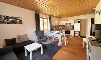 A modern living and dining room with an open kitchen. The decor is simple and cozy, with wooden details and bright light.